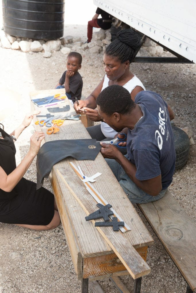Two workers create custom leather goods at Deux Mains in Haiti while a child watches