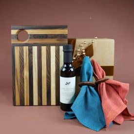 A holiday gift set featuring a mixed wood cutting board, olive oil, sustainably made napkins and wooden napkin rings